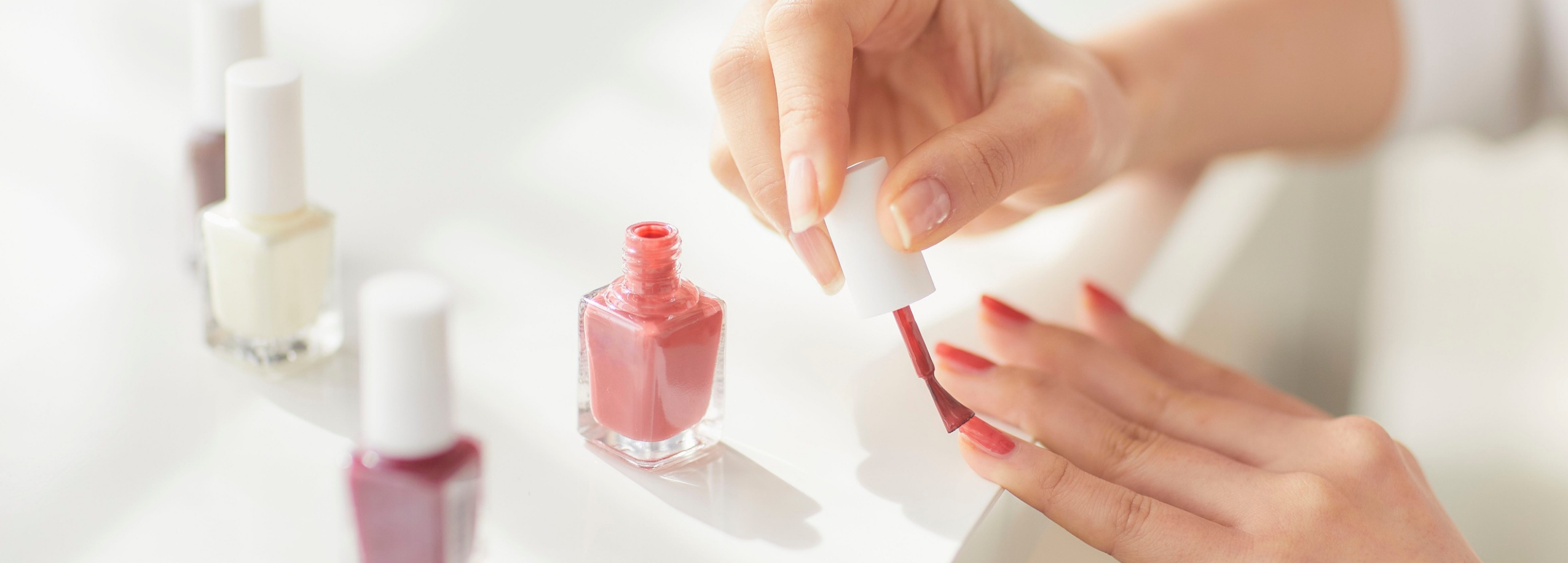 Close-up of a woman applying pink nail polish on her fingers with several other polish bottles blurred in the background, preparing for a DIY manicure.