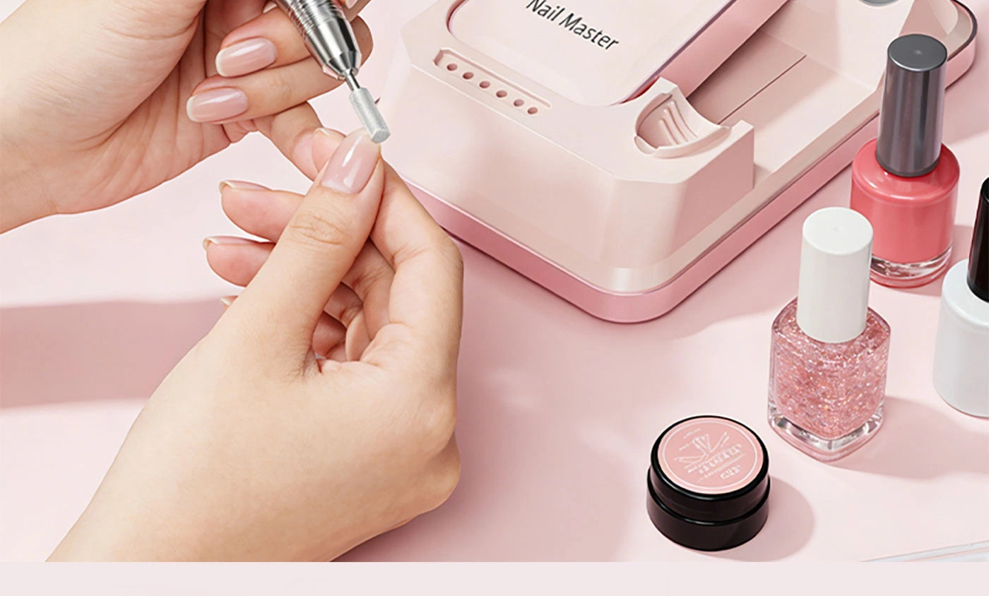 Close-up of a professional cordless e-file smoothly shaping acrylic nails. The pink desktop drill station sits neatly on a clean workspace surrounded by colorful gel polish bottles.