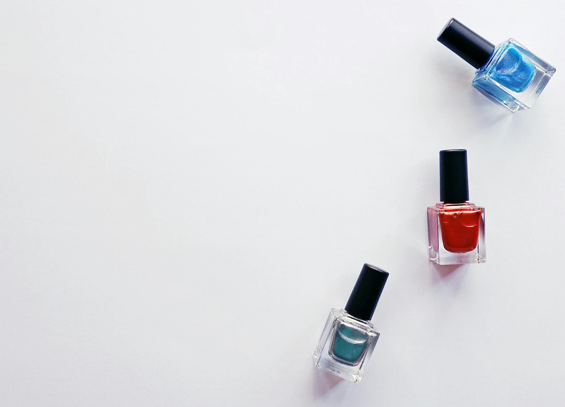 Three glass bottles of shimmering blue, red, and silver nail polish arranged on a white background with negative space.