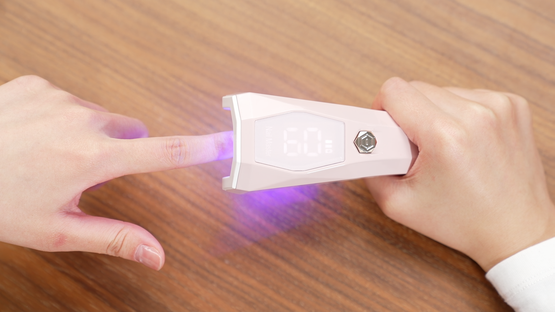 Hands holding a pink rechargeable mini gel nail dryer over one finger on a wooden table. Features a clear LCD screen.